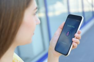 A woman using a voice search feature on her smartphone, with the screen displaying "Speak now" and a microphone icon, illustrating the concept of voice search optimisation. The background is blurred, focusing attention on the interaction with the voice assistant technology.