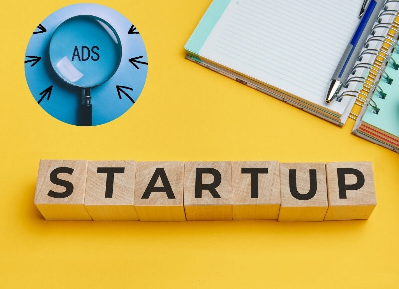 A magnifying glass with "ADS" on it highlights "STARTUP" blocks. Google Ads for startup success, illustrated on a yellow background.