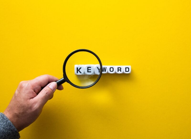 A hand holds a magnifying glass over the word "KEYWORD" on blocks, highlighting the importance of keyword relevance.