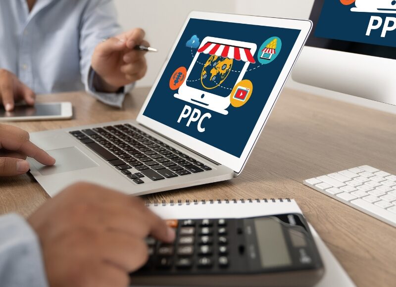 Business professionals analyzing PPC investment timing on a laptop, discussing ad strategies, and using a calculator for budget planning.