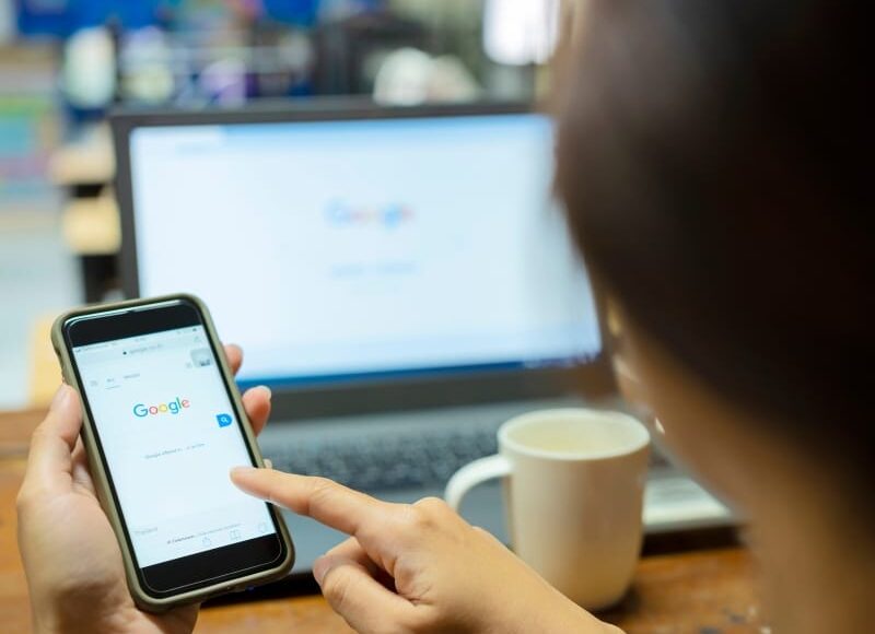 Person using Google on a smartphone while working on a laptop, showing how a google ads service supports online visibility.