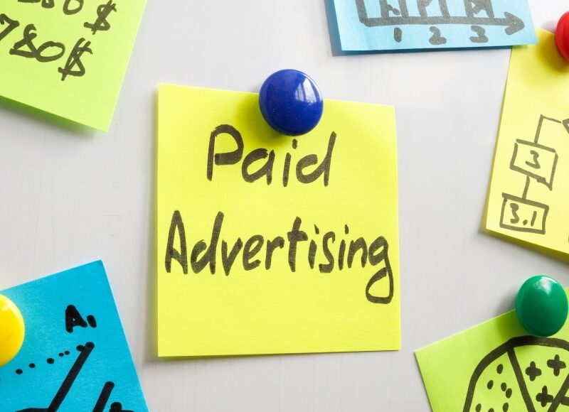 Yellow sticky note on a board with “Paid Advertising” written in marker, used to explain paid Google advertising concepts.